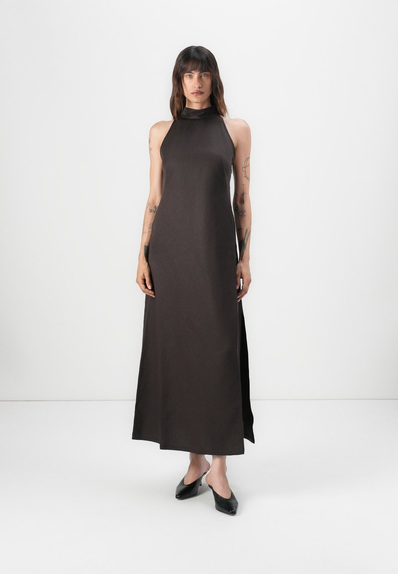 Donkerbruin, mouwloze maxi-jurk met een hoge hals en zijsplitten, gemaakt van een gladde stof. Model draagt zwarte instapschoenen.