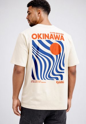 Man wearing cream t-shirt with blue wavy lines, an orange circle, and text "OKINAWA," "Japan the Island of Balance and Harmony," and "Kyūshū" on back.