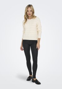 Crème textuurtrui met een ronde hals, gecombineerd met zwarte fitted broek en zwarte instappers, staande tegen een neutrale achtergrond.