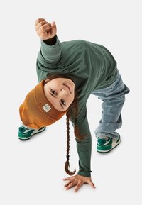 Green long-sleeve shirt, blue jeans, and orange beanie. Child crouching with hand on the ground, displaying green sneakers with white accents.
