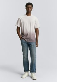 Lichtbeige tot donkergrijs ombre T-shirt, gecombineerd met blauwe rechte jeans en witte sneakers. Het shirt heeft een klassiek crewneck ontwerp.