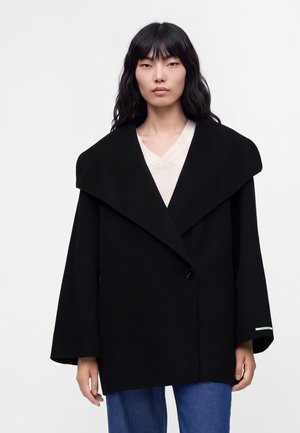 Kvinde med langt sort hår iført en sort oversized frakke med bred krave og blå jeans mod en ensfarvet lys baggrund.