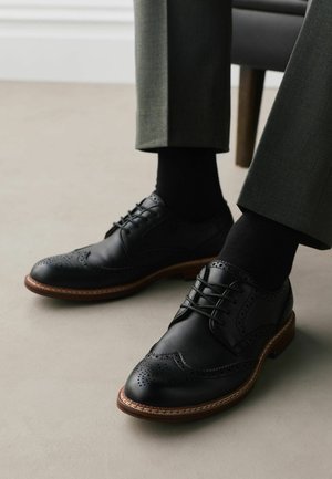 Black leather brogue shoes with brown soles worn with black socks and dark gray trousers seated on a chair.