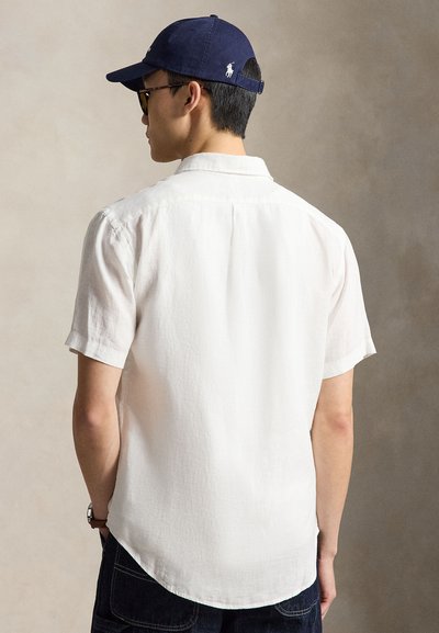 Muž oblečený v bielu košeľu s krátkym rukávom a námorníckou modrou čiapkou, stojaci chrbtom ku jednolitému béžovému pozadiu.