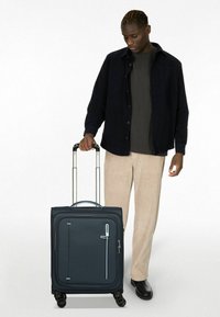 Valise à roulettes en tissu bleu foncé avec fermeture éclair, poignée supérieure, poche avant et quatre roues ; le modèle porte une chemise noire et un pantalon beige.