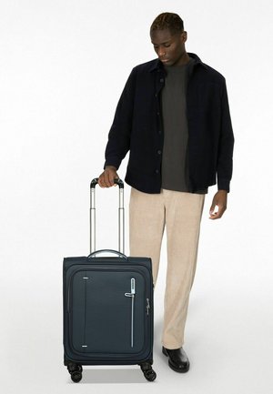 Rolling-Trolley aus dunkelblauem Stoff mit Reißverschluss, Tragegriff, Fronttasche und vier Rädern; Modell trägt ein schwarzes Shirt und beige Hose.