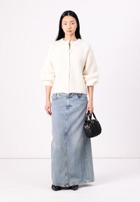 Krémový rebrovaný kardigán s gombíkovým zapínaním v kombinácii s dlhou modrou riflovou sukňou. Modelka drží malú čiernu kabelku. Hladké textúry a ležérny dizajn.