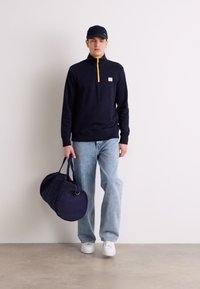 Pulover navy cu fermoar galben, blugi albaștri deschiși, adidași albi și o geantă de voiaj navy în mână. Purta o șapcă navy.