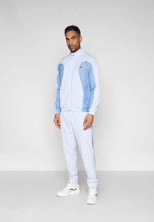 Lichtblauw trainingspak met donkerblauwe accenten, ritsjas en elastische manchetten. Gecombineerd met witte sportschoenen. Gladde textuur.