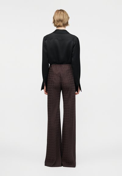 Άτομο με κοντά ξανθά μαλλιά, που φοράει μαύρο μακρυμάνικο τοπ και καφέ σκούρο παντελόνι με φαρδιά πόδια και ψηλή μέση, εμφανίζεται από πίσω.