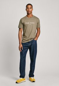 Olivově zelené tričko s logem "Pepe Jeans", kombinované s tmavě modrými džíny a žlutými teniskami. Jednoduchý design, kulatý výstřih, volný střih.