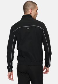 Zwarte windjack met witte accenten, voorzien van een hoge kraag, elastische manchetten en een trekkoord onderaan. Logo op de rug weergegeven.