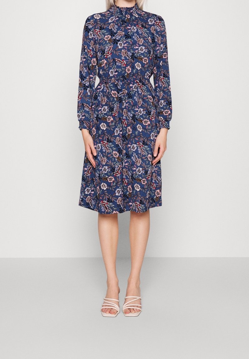 Femme portant une robe fleurie bleu marine longue jusqu'aux genoux, à manches longues et col montant, assortie à des talons blancs ouverts à brides.