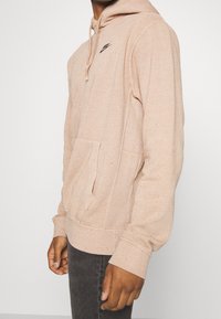 Ljusrosa hoodie i texturerad tyg, med en framficka och svart logotyp på bröstet.