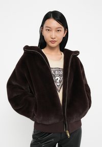 Veste zippée en fausse fourrure marron avec capuche. Présentant un ourlet et des poignets côtelés contrastants, ainsi que des poches latérales. Portée par-dessus un t-shirt graphique beige.