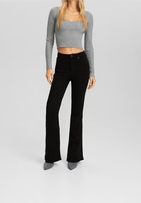 Mujer vistiendo un top corto de manga larga gris con ribetes, jeans negros de talle alto acampanados y zapatos grises de punta, de pie sobre un fondo liso.