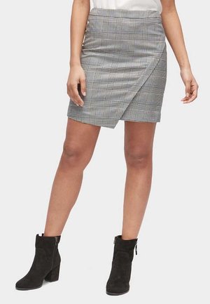 Kobieta w szarej spódnicy w kratę, wiązanej na zakładkę, zestawionej z czarnymi zamszowymi botkami na kostkę na jednolitym tle.