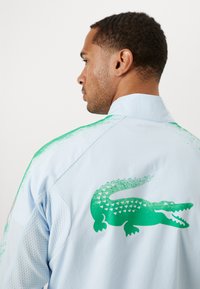Jachetă de culoare albastru deschis, cu un logo de crocodil verde pe spate, realizată dintr-un material texturat și cu accente în degrade în zona umerilor.