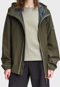 Personne portant une veste de pluie à capuche vert olive ouverte par-dessus un t-shirt beige clair et un pantalon foncé, debout devant un fond uni.
