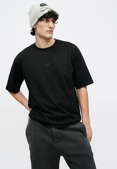 Black cotton t-shirt with short sleeves and embossed logo on the chest, paired with dark gray pants and a light gray beanie featuring a face pattern.