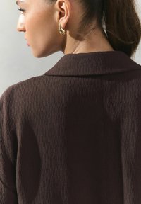 Brauner, strukturierter Stoffjacke mit Kragen, die sanfte Wellen und eine lockere Passform aufweist. Goldene Creolen sind im Profil sichtbar.