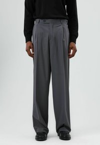 Pantalon gris à jambes larges avec un devant plat, une fermeture à un seul bouton et des poches latérales. Tissu lisse avec une légère brillance, associé à des chaussures noires.