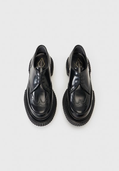 Chaussures derby en cuir noir poli avec des détails en brogue et des semelles épaisses à rainures, vues de dessus sur un fond clair.