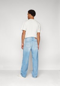Jeans de mezclilla anchos de color azul claro con un acabado deslavado, detalles de bolsillos y una camiseta de algodón blanca. Mostrados desde atrás. Zapatillas blancas.