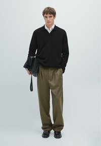 Black knitted sweater with a V-neck, olive green pleated trousers, and glossy black shoes. Model holds a black leather bag.