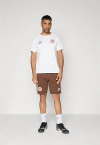 Hvid atletisk t-shirt med logo, kombineret med brune shorts med sidestriber og logo. Sorte atletiksko og hvide sokker fuldender looket.