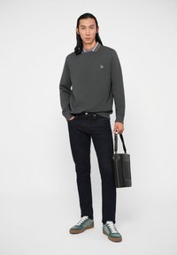 Pull gris avec un design en V, jeans foncés et baskets teal. Tenant un sac noir avec un détail texturé et une bandoulière. Col rayé simple visible.