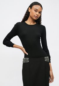 Žena s dugom kovrčavom kosom koja nosi uski crni rebrasti top i crnu suknju s ukrasima od srebrnog lanca na bokovima, pozira s rukom na struku.