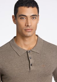 Brązowa koszulka polo wykonana z dzianiny, z klasycznym kołnierzykiem, trzema ciemnymi guzikami oraz subtelnie wyhaftowanym logo na piersi.