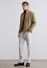 Γούνινο τζάκετ ταμπά σουέντ με ριμπ πλέξη και ψηλό γιακά, φορεμένο πάνω από καφέ πουλόβερ, συνδυασμένο με λευκά τζιν και μαύρα μοκασίνια.