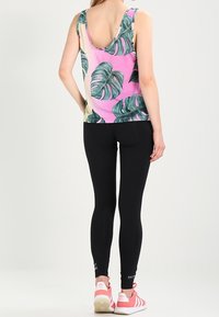 Kvinna klädd i en ärmlös rosa topp med gröna bladmönster, svarta leggings och rosa Adidas-sneakers, stående med ryggen mot kameran.