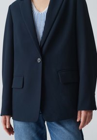 Blazer bleu marine avec deux poches avant, fermeture à un bouton et revers crantés, associé à un haut rayé clair et un jean bleu.