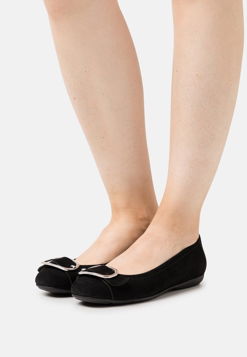Paire de chaussures plates noires avec détail de boucle argentée portées pieds nus, présentées sur fond blanc.