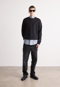 Sweatshirt preto com logótipo "Boss" em relevo, camisa colarinho azul claro por baixo, jeans pretos e grandes óculos de sol quadrados pretos.