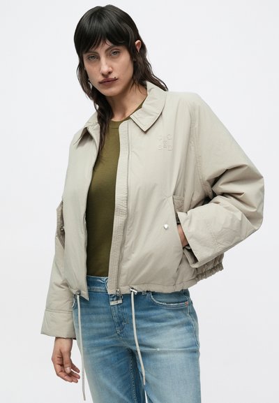 Μπεζ cropped μπουφάν bomber με φερμουάρ στο μπροστινό μέρος, πλαϊνές τσέπες και σκοινάκι στο τελείωμα· διαθέτει γιακά και απαλό, ελαφρύ ύφασμα.