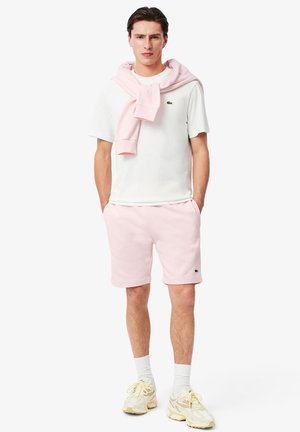 T-shirt blanche, shorts roses, pull rose drapé autour des épaules, logo noir sur la poitrine gauche, chaussettes blanches, baskets claires.
