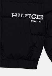 Černá prošívaná bunda s přední kapsou a bílým logem "HILFIGER NEW YORK". Tkanina vypadá hladce s jemným lesklým povrchem.