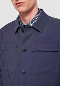 Giacca blu navy con due tasche sul petto, chiusura con bottoni e design con colletto. Il materiale sembra liscio con sottili dettagli di cucitura.