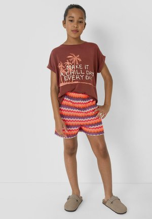 Young girl wearing a brown t-shirt with palm trees and text, colorful zigzag shorts, and beige slip-on shoes, standing with one hand on hip.