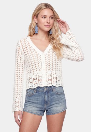 Ung kvinde med langt blondt hår iført blå frynseøreringe, en hvid hæklet langærmet top og denimshorts, stående mod en hvid baggrund.
