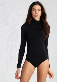 Γυναίκα με μακριά καστανά μαλλιά που φοράει ένα εφαρμοστό μαύρο μπουφάν με μακριά μανίκια και λαιμόκοψη εναντίον ενός απλού ανοιχτού φόντου.