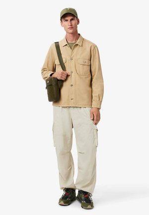 Giacca marrone con tasche frontali, camicia verde chiaro, pantaloni cargo beige e scarpe nere con dettagli rossi. Tiene una borsa verde oliva.