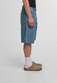 Blå denimshorts med lige snit og knælangt design, parret med beige ruskindsklogs og hvide ankelsokker.