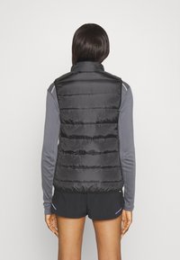 Svart puffväst med hög krage, horisontell quiltning och slät satinfinish. Bärs över en långärmad grå skjorta och svarta shorts.