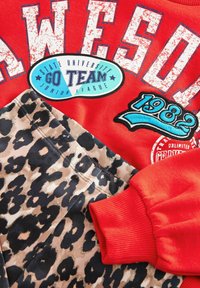 Sweatshirt rouge avec le texte "AWESOME", orné de patchs bleus et noirs. Complété par un pantalon beige avec un motif de léopard noir.
