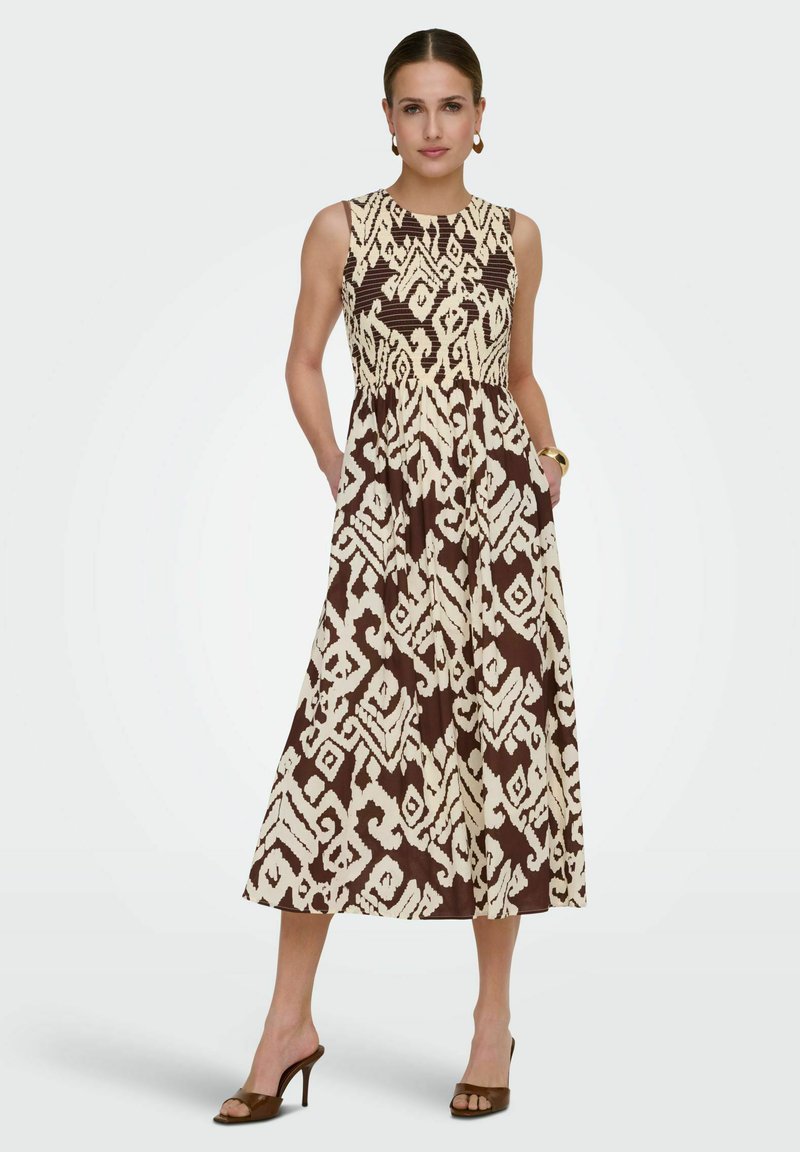 Femme portant une robe midi sans manches à motifs marron et beige avec poches, sandales marron à talons, boucles d'oreilles créoles dorées et bracelet, debout.